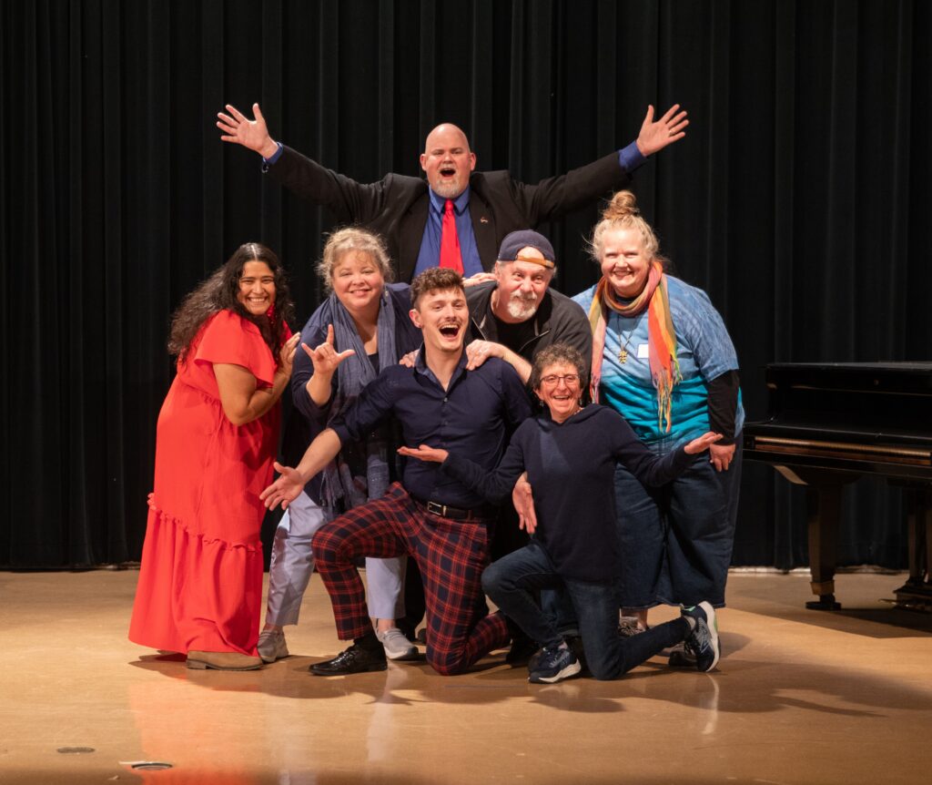 Seven people pose exuberantly on a stage in front of a black curtain; one stands tall with arms raised wide, others crouch and lean in, laughing and gesturing playfully.