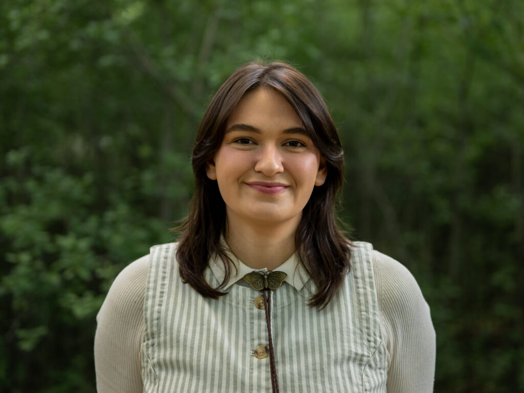 Headshot of a person with shoulder-length dark brown hair and curtain bangs, wearing a light green and white striped vest over a cream long-sleeve shirt, with a bronze moth bolo tie. They smile warmly against a blurred green tree background.