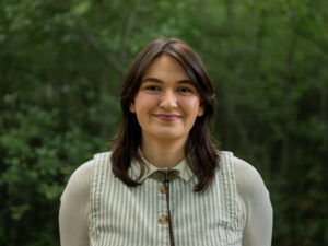 Headshot of a person with shoulder-length dark brown hair and curtain bangs, wearing a light green and white striped vest over a cream long-sleeve shirt, with a bronze moth bolo tie. They smile warmly against a blurred green tree background.