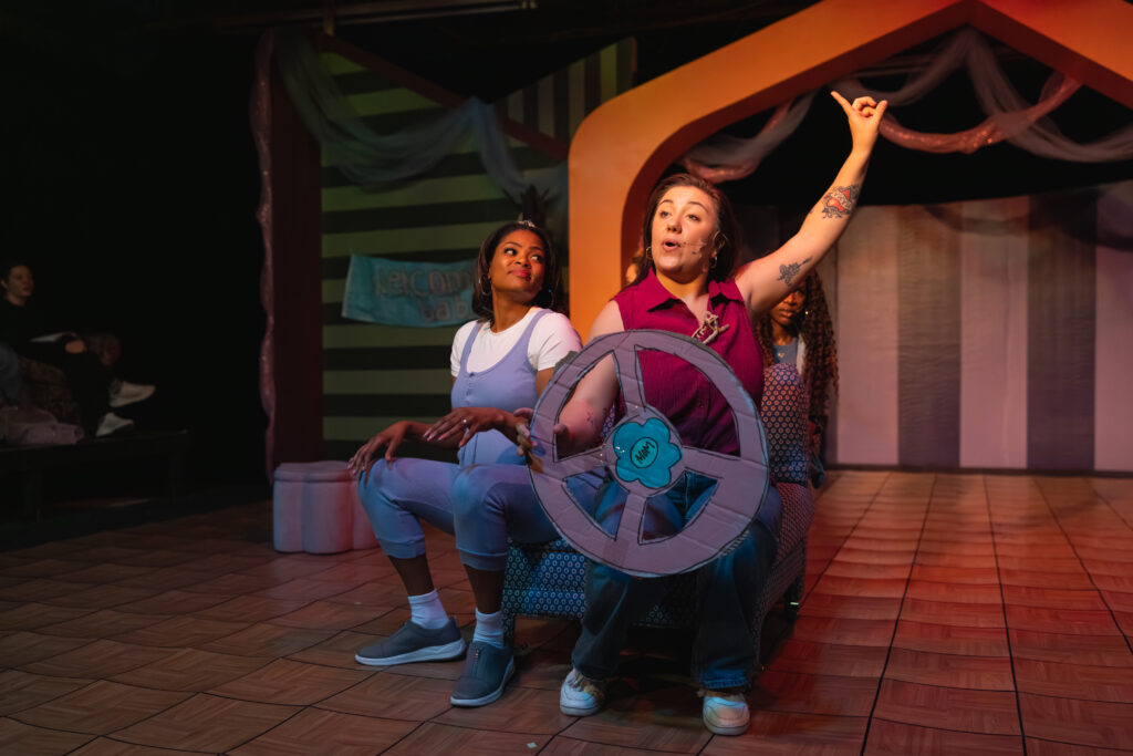 two women sitting on a performance stage, with one holding a cardboard steering wheel and pointing in the air