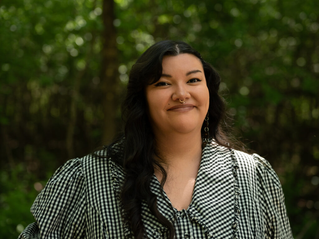 Headshot of a person with long, wavy black hair, wearing a black and white gingham top with ruffle detailing, gold hoop earrings, and a small nose ring. They smile warmly against a blurred green forest background.