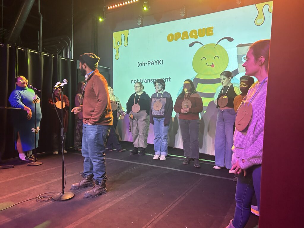 Participants wearing numbered medallions stand on a stage in front of a projected bee-themed spelling bee slide reading "OPAQUE. Not transparent." A host stands at the microphone and an emcee stands at a podium to the left.