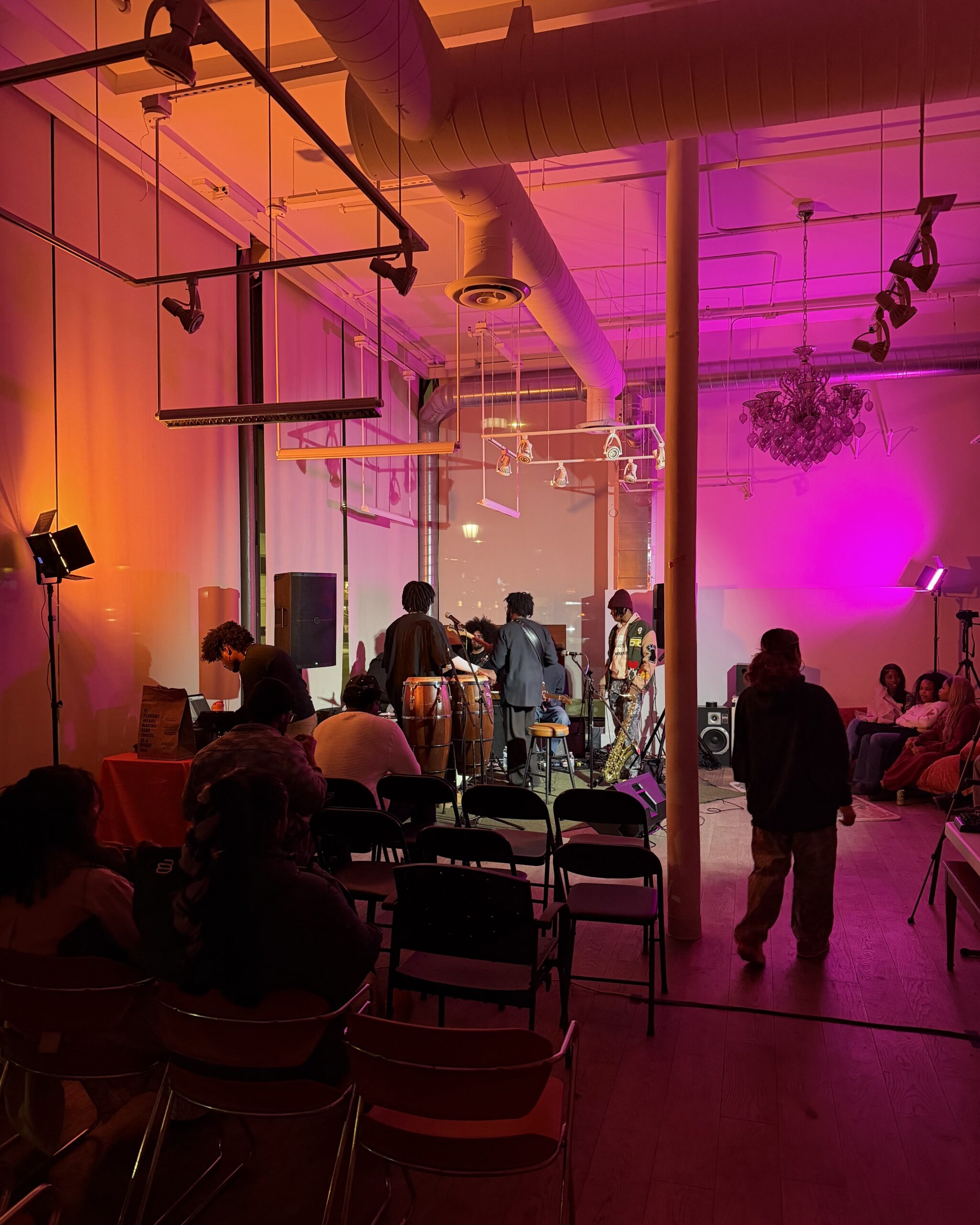 Four people stand on a makeshift stage in the corner of ZOMA House. The space is dark and lit with pink and orange flood lights.