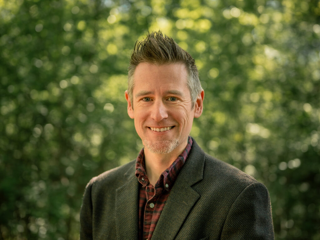 Headshot of a person with salt-and-pepper hair styled upward and light stubble, wearing a dark textured blazer over a plaid shirt. They smile warmly against a blurred bright green tree background.