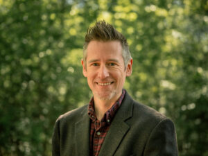 Headshot of a person with salt-and-pepper hair styled upward and light stubble, wearing a dark textured blazer over a plaid shirt. They smile warmly against a blurred bright green tree background.