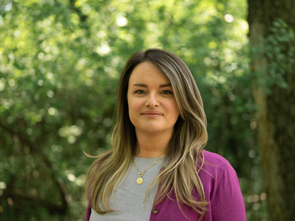 Headshot of a person with long, wavy ombre hair transitioning from dark brown roots to lighter ends, wearing a magenta cardigan over a striped shirt and a gold pendant necklace. They smile softly against a blurred green tree background.