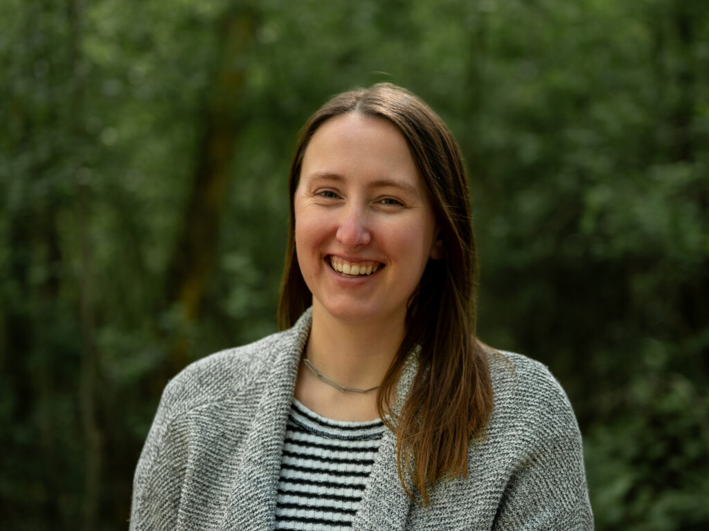 Headshot of a person with long, straight light brown hair, wearing a gray knit cardigan over a striped shirt and a thin silver necklace. They smile broadly against a blurred green tree background.