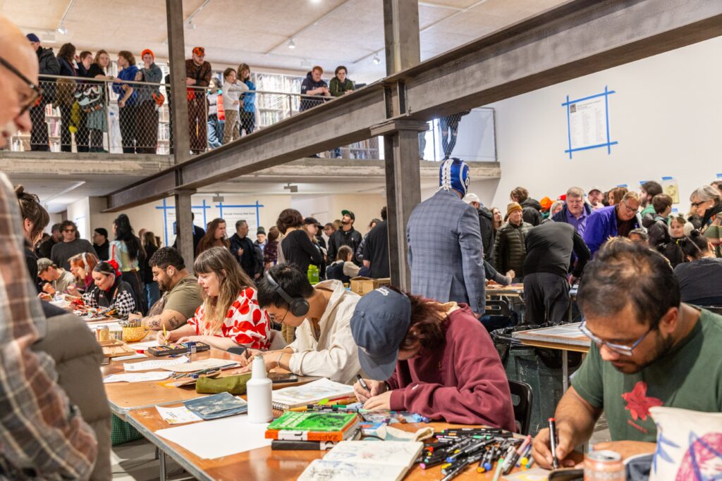 A packed two-story gallery space buzzes with energy as dozens of people draw at long tables crowded with markers and sketchbooks, while more spectators look on from a mezzanine level above.