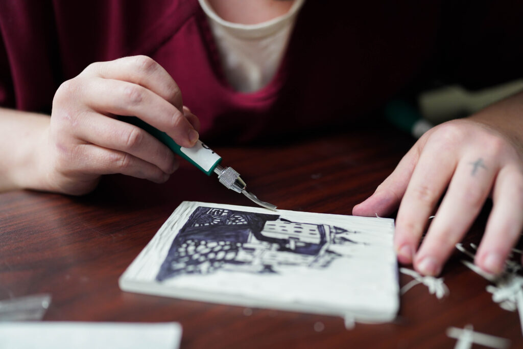 A person using a carving tool to cut a seaside castle design into a printing block.