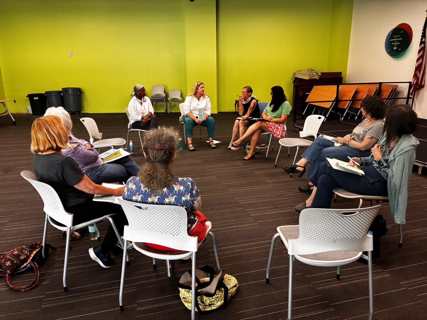 Alison Pelegrin and Khadijah Queen face a circle of about ten seated participants in a bright yellow-green room, facilitating a community workshop discussion. Several participants hold papers and notebooks.