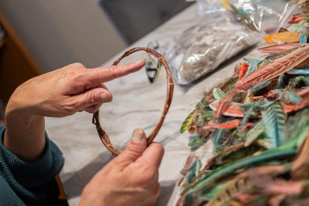 One medium-light skin tone hand points at a detail of a dreamcatcher in progress, while the other hand holds the hoop. There are brightly colored feathers piled to the right.