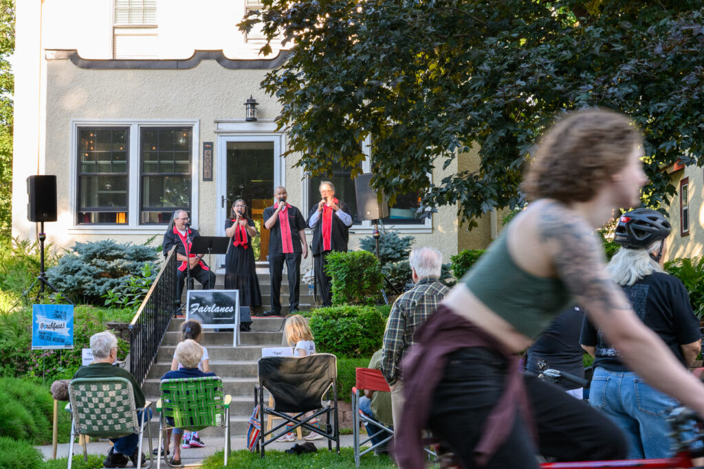 The Fairlanes, a four-person vocal group dressed in black with red sashes, perform on the front steps of a stucco house. Audience members watch from lawn chairs in the foreground while a cyclist passes in soft focus at right.