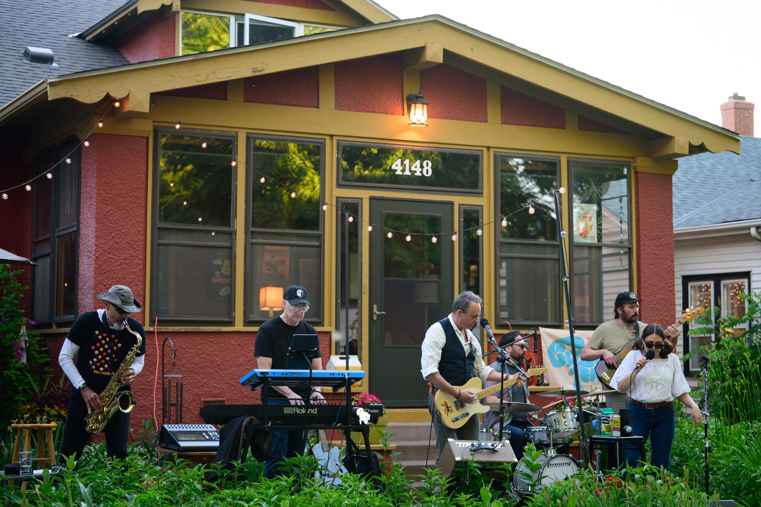 A six-piece band performs in the front garden of the red craftsman bungalow at 4148 as dusk settles