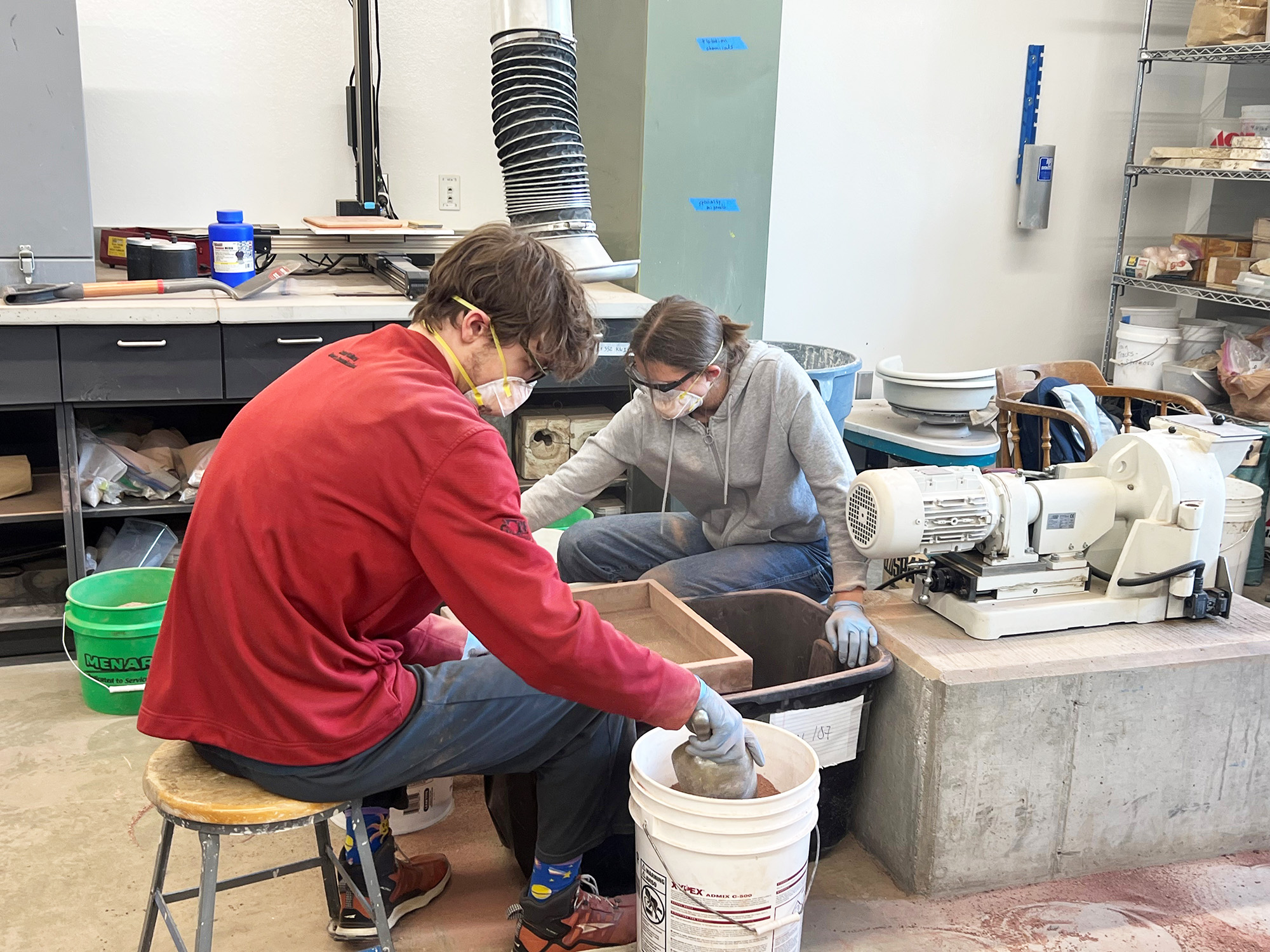 Two people wearing masks and gloves, processing clay indoors with large buckets.