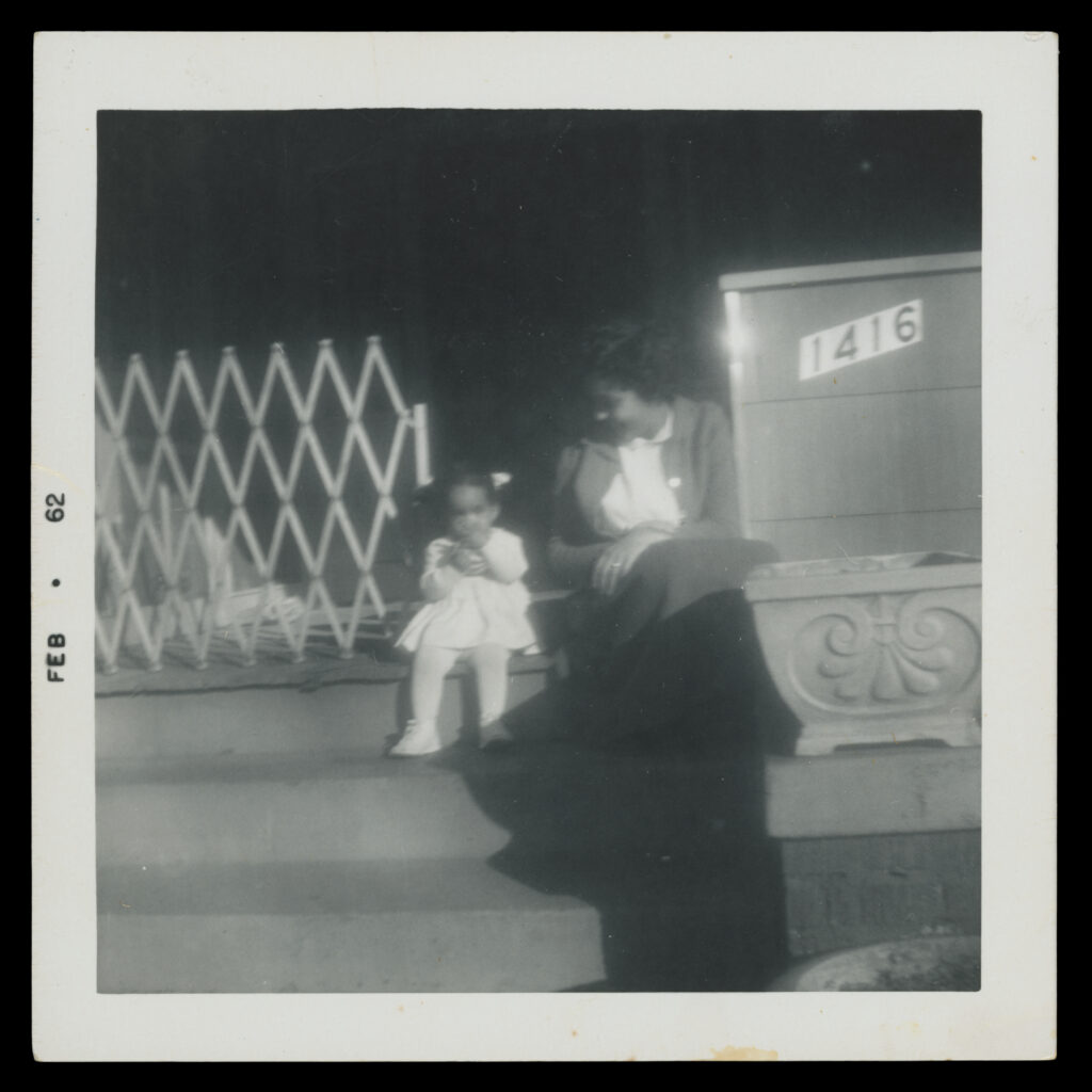 A black and white archival photo of an adult and young child sitting on the porch stairs of a house