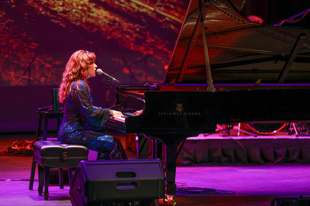 A performer with light skin tone and long red hair sings and plays a Steinway & Sons grand piano on a stage bathed in pink and red light, wearing a sparkling blue gown.