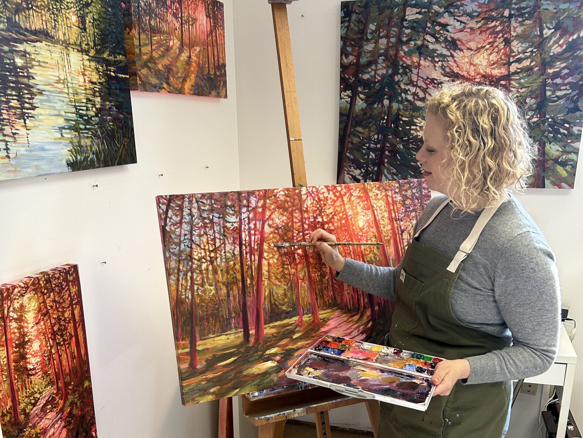 A woman with light skin and blonde hair painting a treed landscape in a studio.