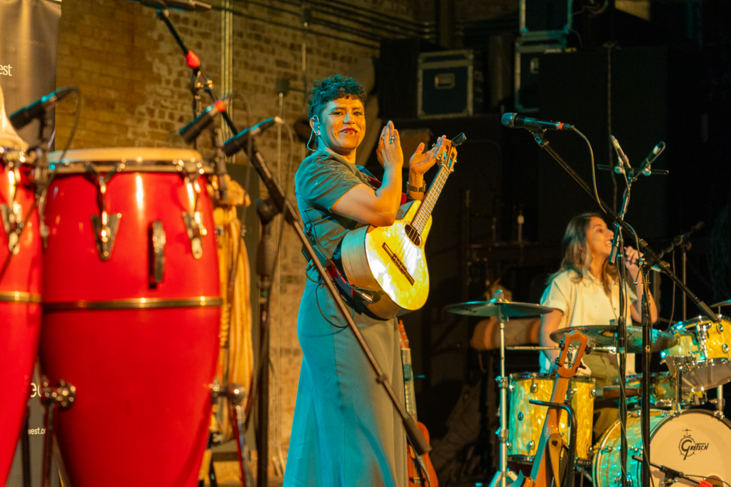 A woman wearing a bandola (a small string instrument like a guitar) looks directly to camera with her hands raised to clap, as a pair of congas sit to her right and a drummer behind a drum kit sits to her left.
