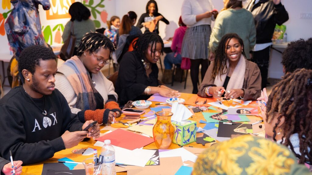A group of people with dark skin tone sit around a table filled with construction paper to create cut paper portraits at ZOMA House.
