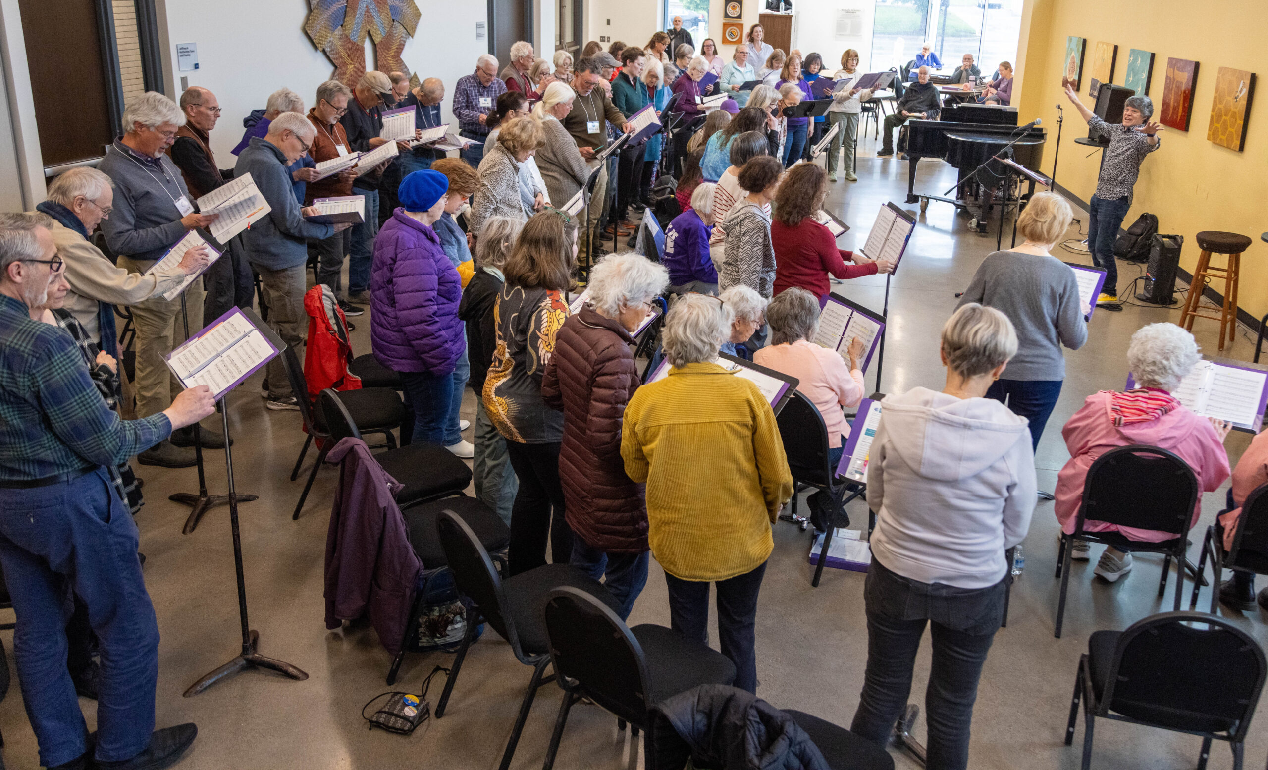 A large intergenerational choir stands and sings together in a rehearsal room.