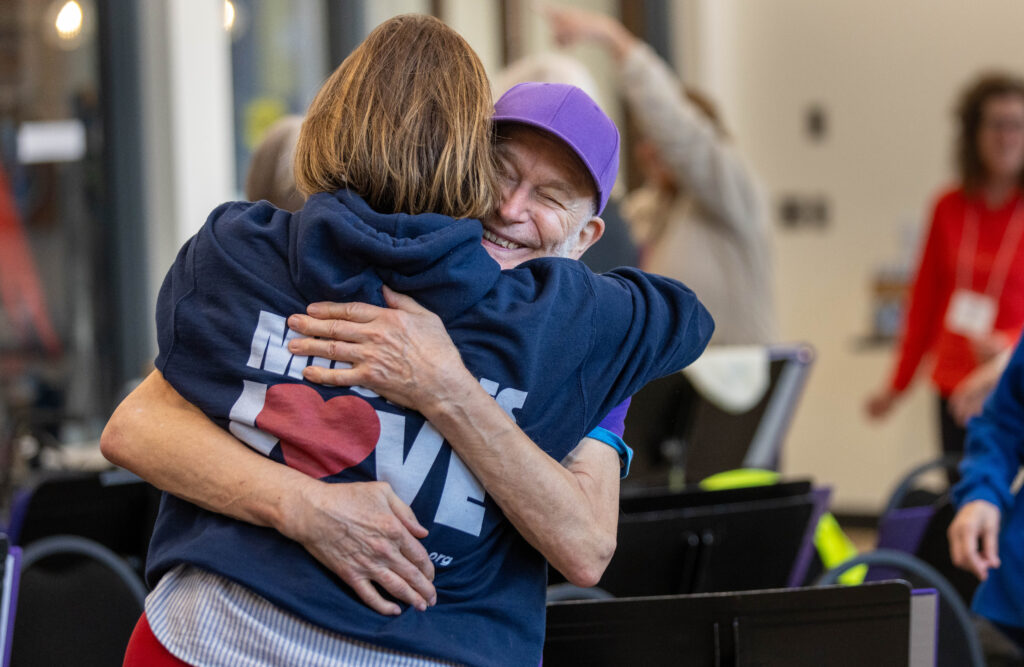 Two older adults embrace warmly at a rehearsal; one wears a purple cap and grins widely, eyes closed, as they hug.