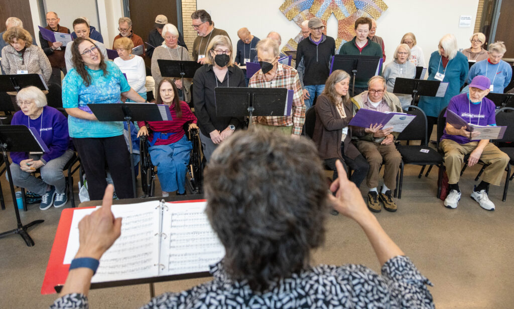 Conductor leads a large intergenerational choir reading from purple music folders.