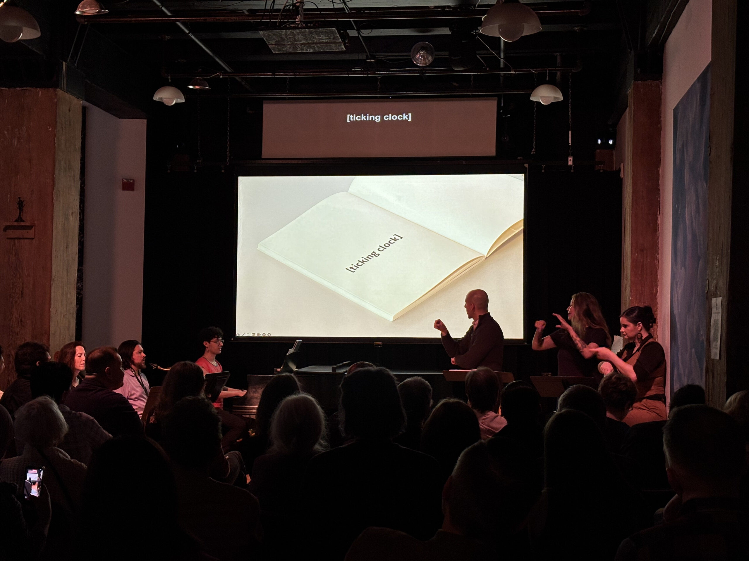 Music-theater performance featuring double screen projections, two narrators, one sign language interpreter, and three musicians. The projection shows a blank book page turned with the caption [ticking clock]. The audience is seen watching attentively.