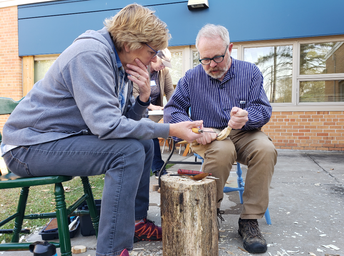 An older man with gray hair and light skin carving a spoon while a woman looks on, holding her tool.