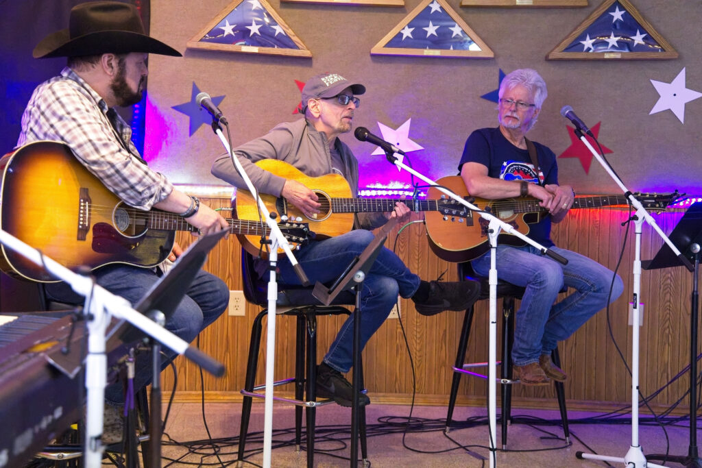 Three people with light skin sitting on stools holding guitars in front of microphones. Folded American flags are in the background.