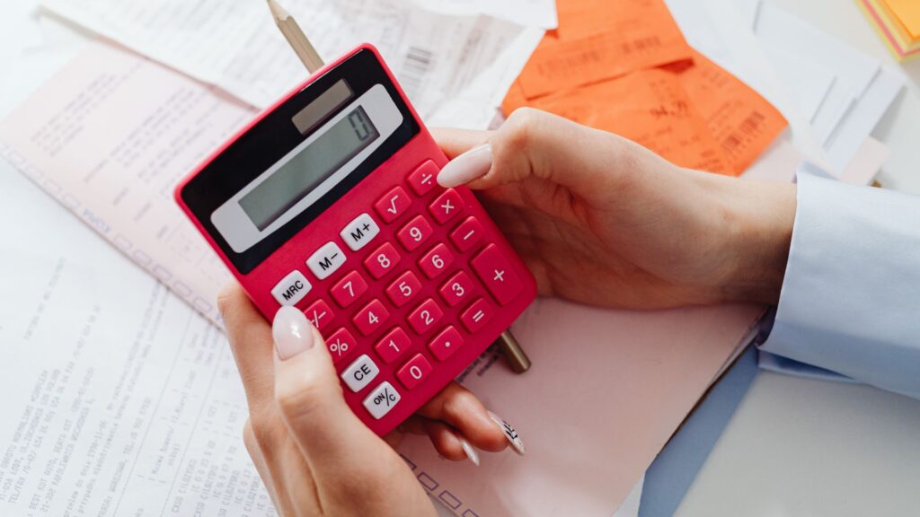 A person with light skin holds a pink calculator atop a pile of papers.