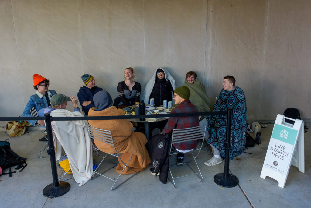 A group of people sitting around a round table. Some have blankets wrapped around them.
