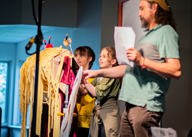 People on a stage holding a large puppet and a script.
