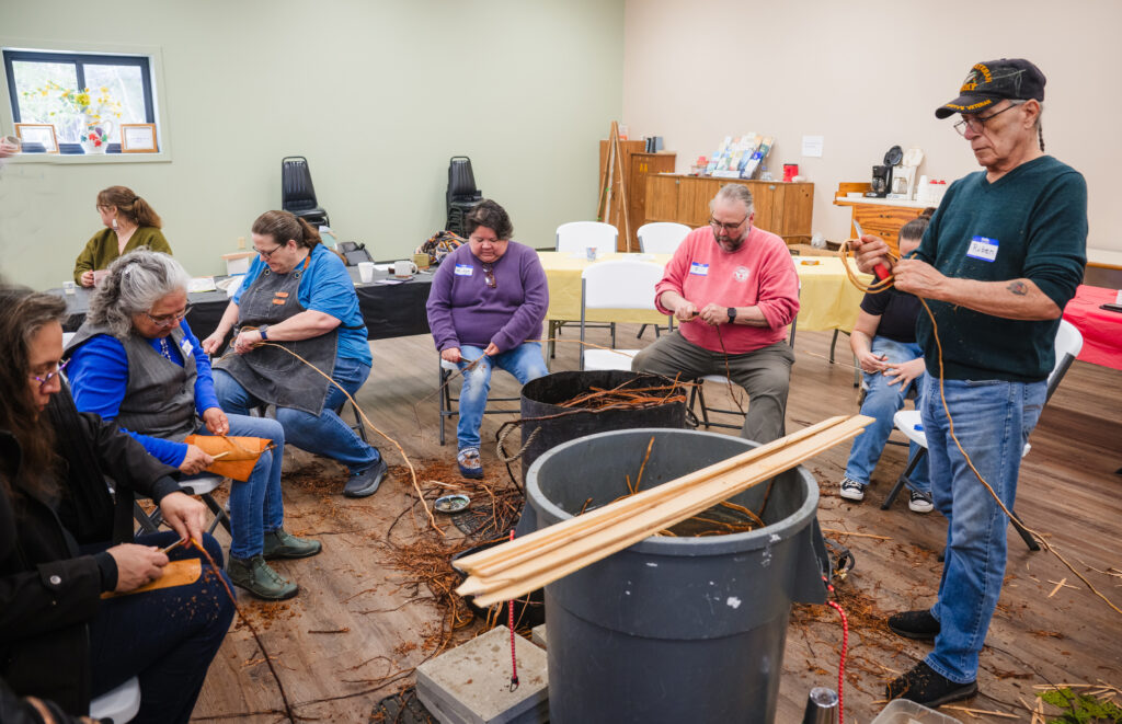 People sitting on chairs in a semi-circle while stripping tree root. There are large buckets by them filled with roots soaked in water.
