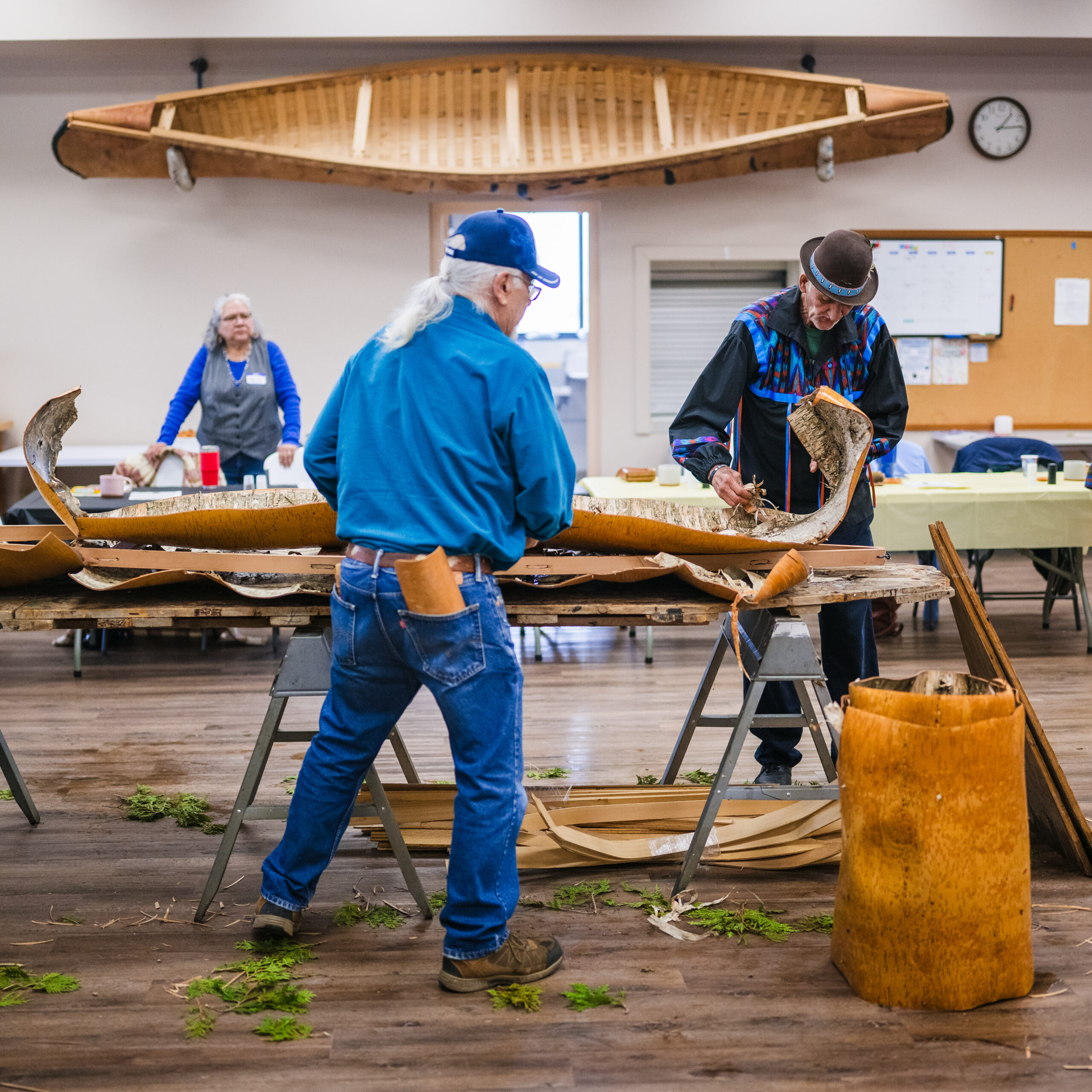 Two people standing by a table as they unroll and layer birch bark in preparation to build a canoe