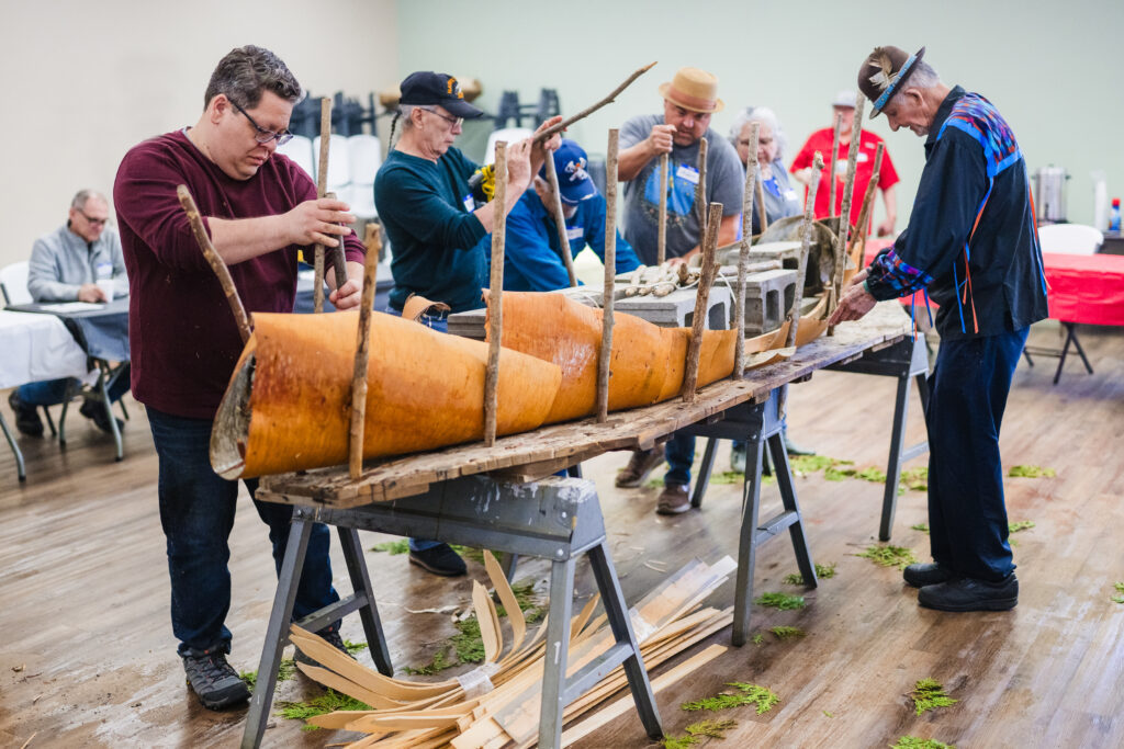A group of people creating a canoe frame on a table using sapling trunks, cinder blocks, and birch bark