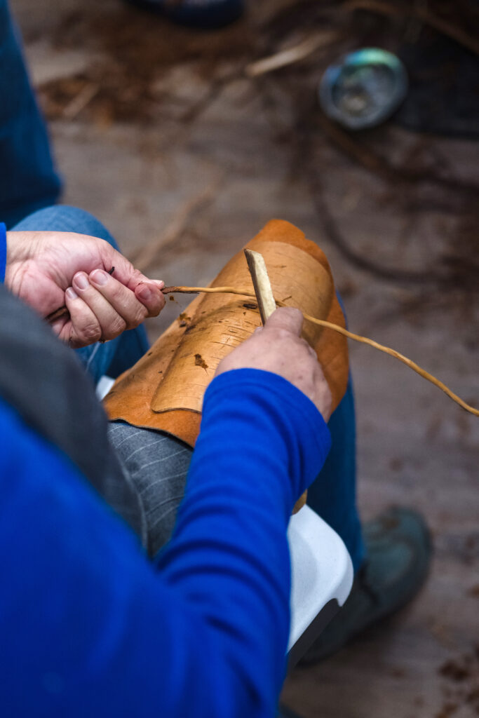 A person stripping a tree root over their right thigh with a tool in their hand