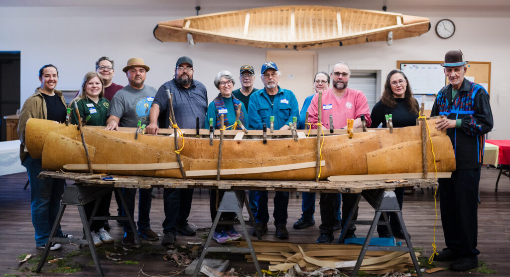 A group of people standing behind an in-progress birch bark canoe build