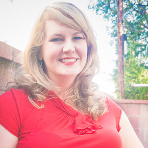Outdoor headshot of a smiling person with light skin tone and long blonde hair, wearing a red shirt and red lipstick.