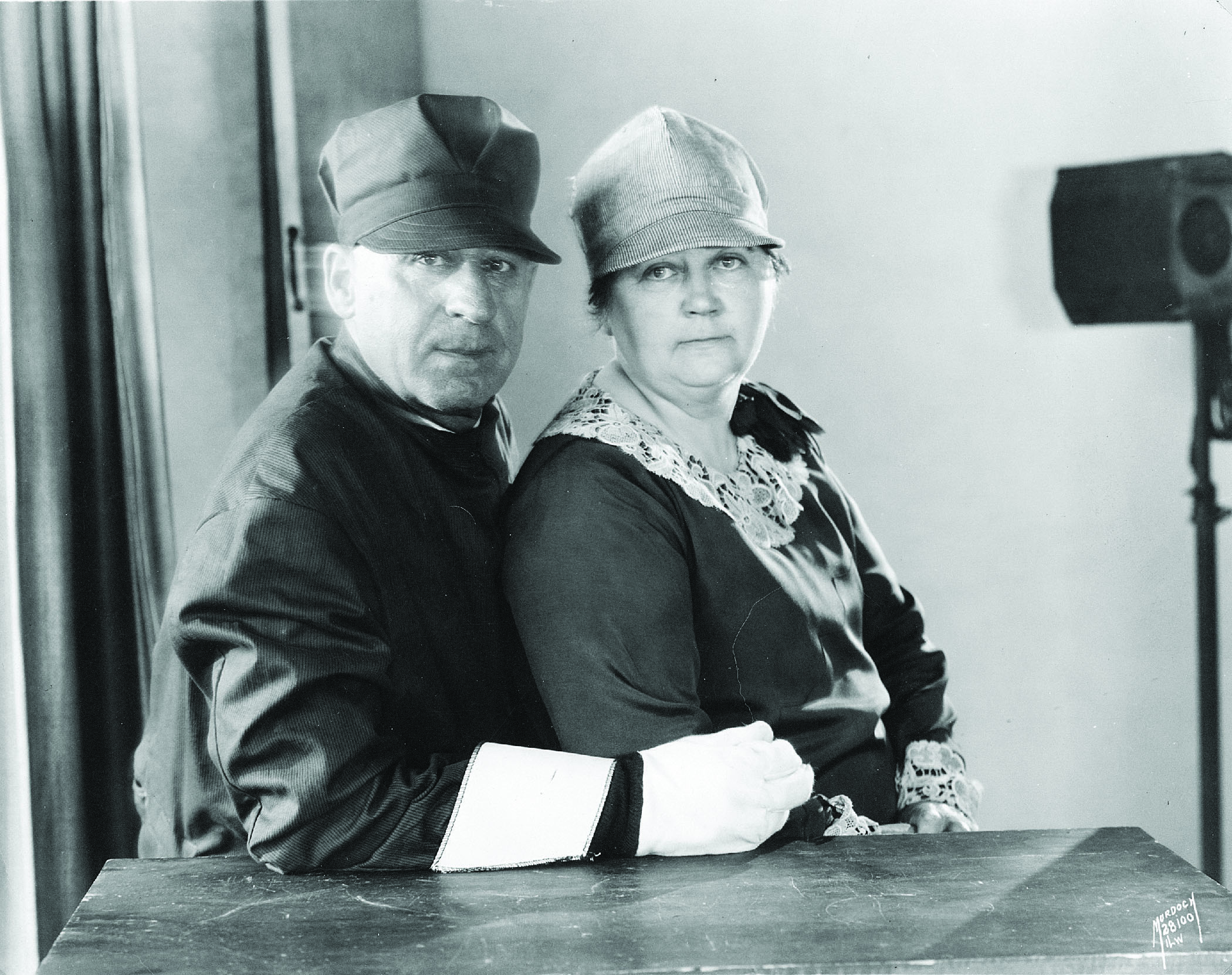 A black and white archival photo of two people sitting close together. Both of them are wearing brimmed caps.
