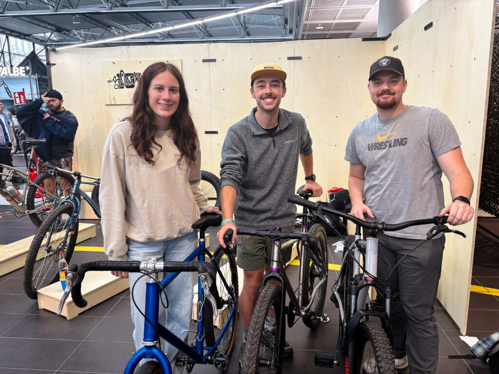 Three people smiling and standing together. Each of them are holding a bicycle by their side.