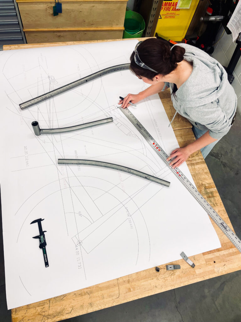 A person holding a long ruler and writing tool on a table as they measure out a diagram of a bicycle printed on paper.