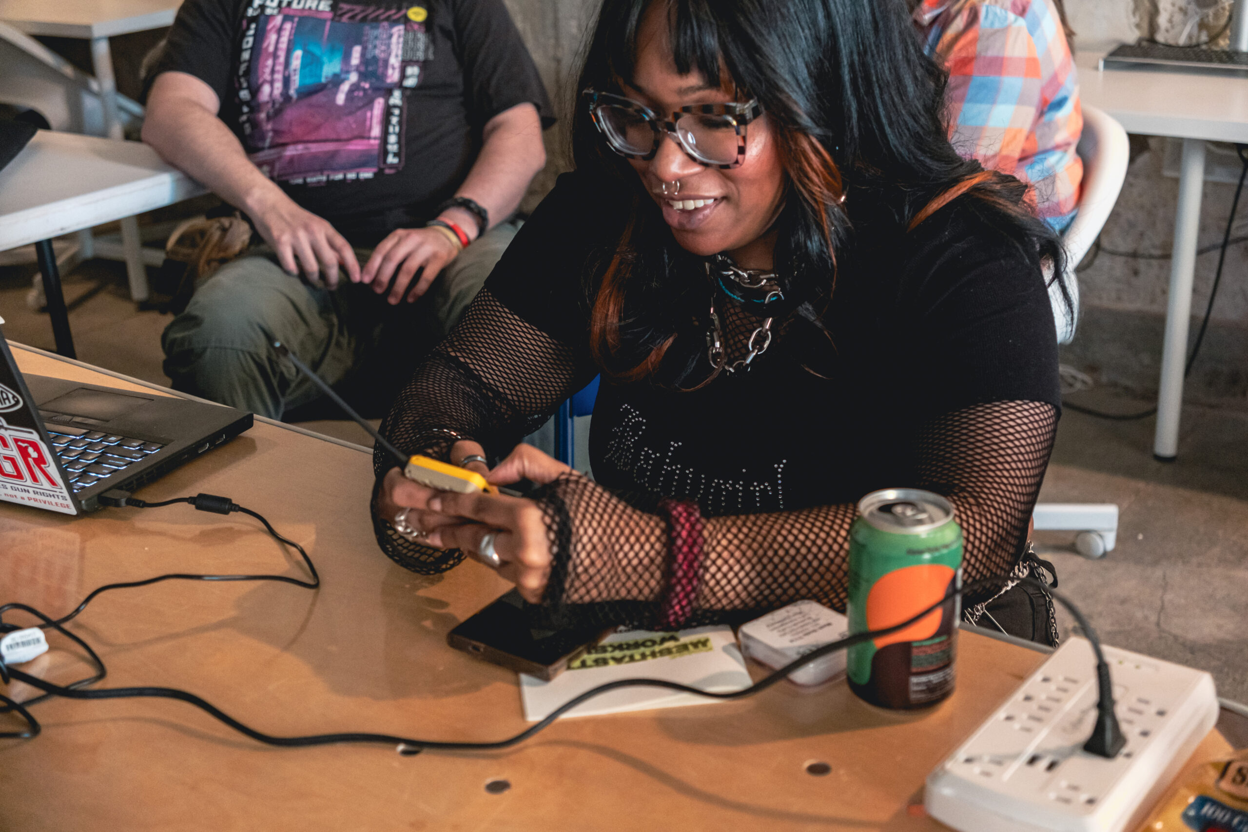 A person with dark skin tone smiling at a table and holding a small electronic device.