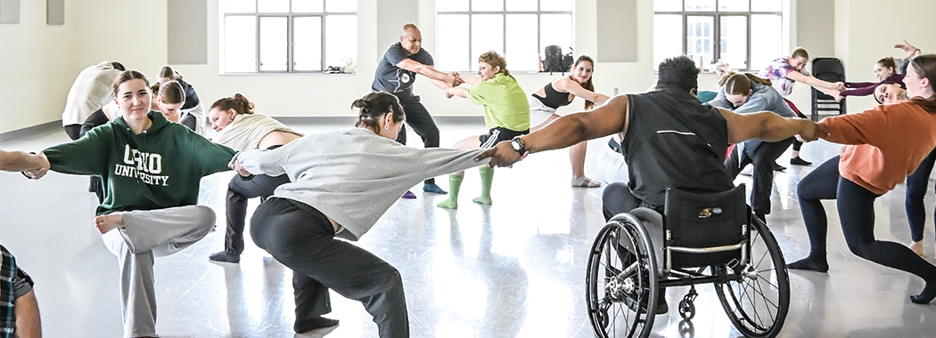 Several dancers fill a brightly lit studio. Most stand in a squat position, and one sits in a wheelchair, as they hold hands with outstretched arms across the studio.