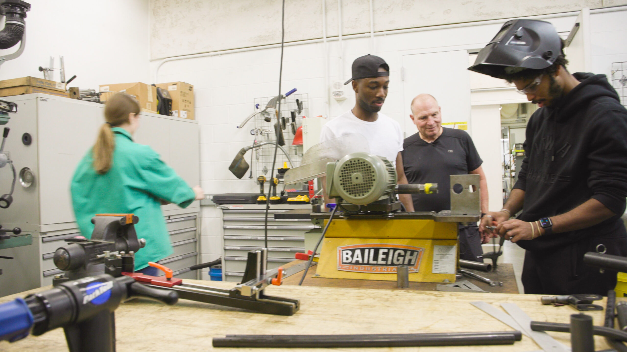 Two people working with a power tool placed on a table as another person looks on