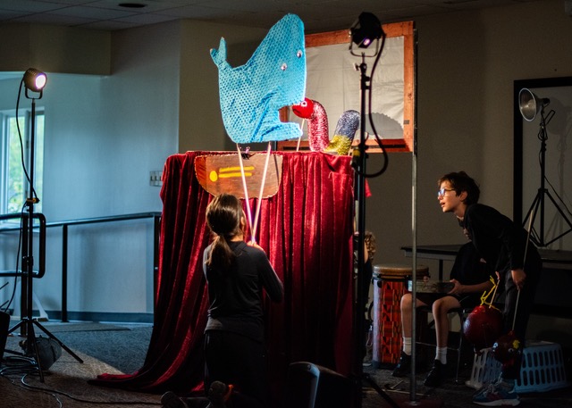 People performing a puppet show on stage with a large blue whale character with googly eyes.