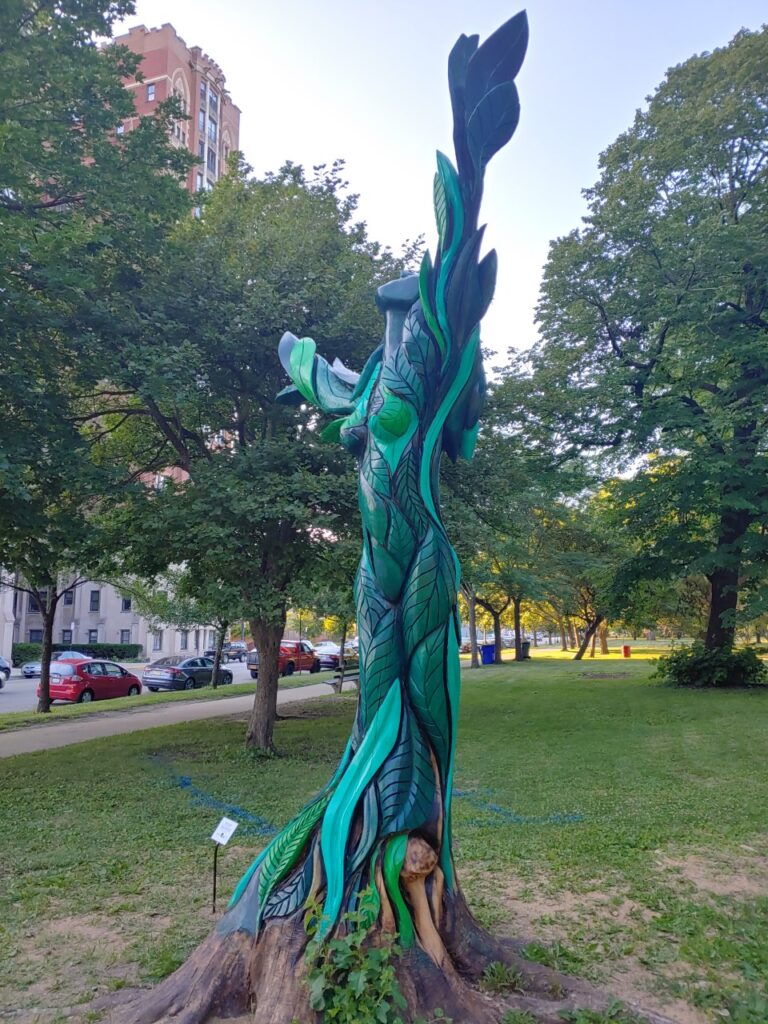 A large green woman carved out of a tree stump on a lawn.