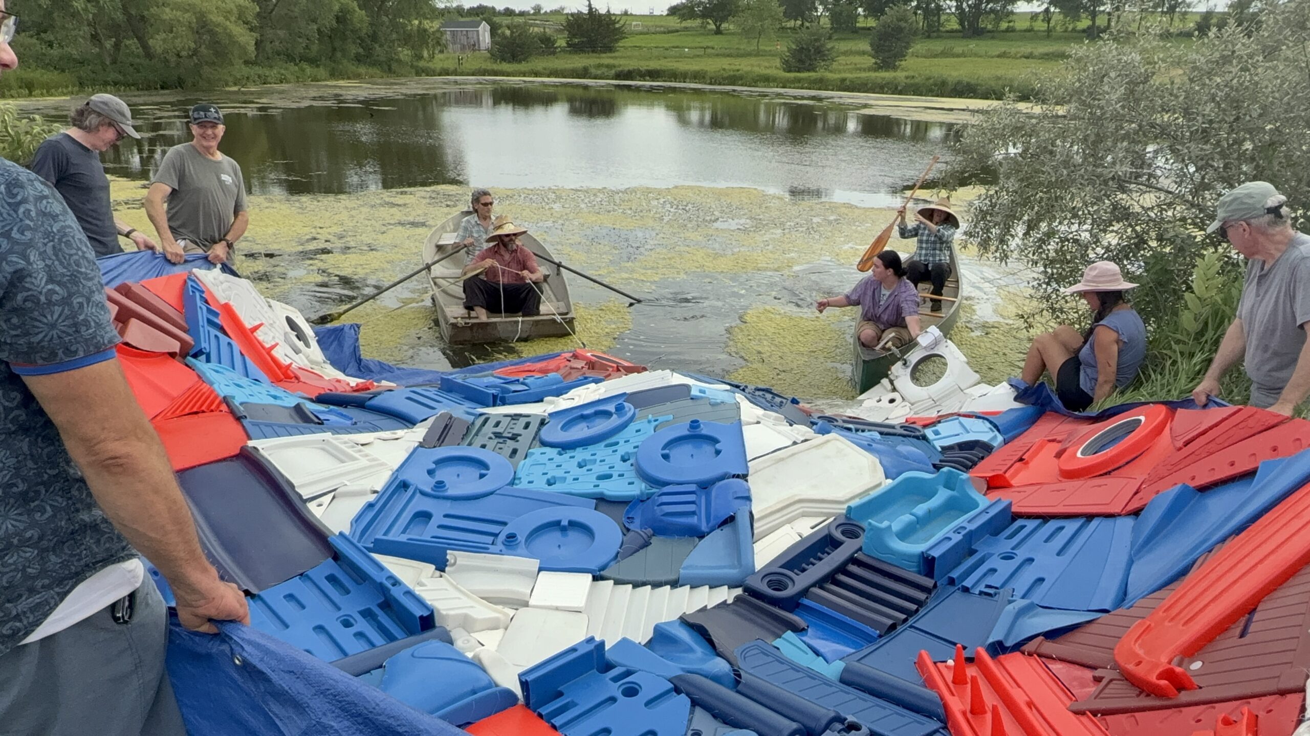 People in a paddle boat and canoe pulling a large sculpture into a pond as others help lift it off the ground by the shore