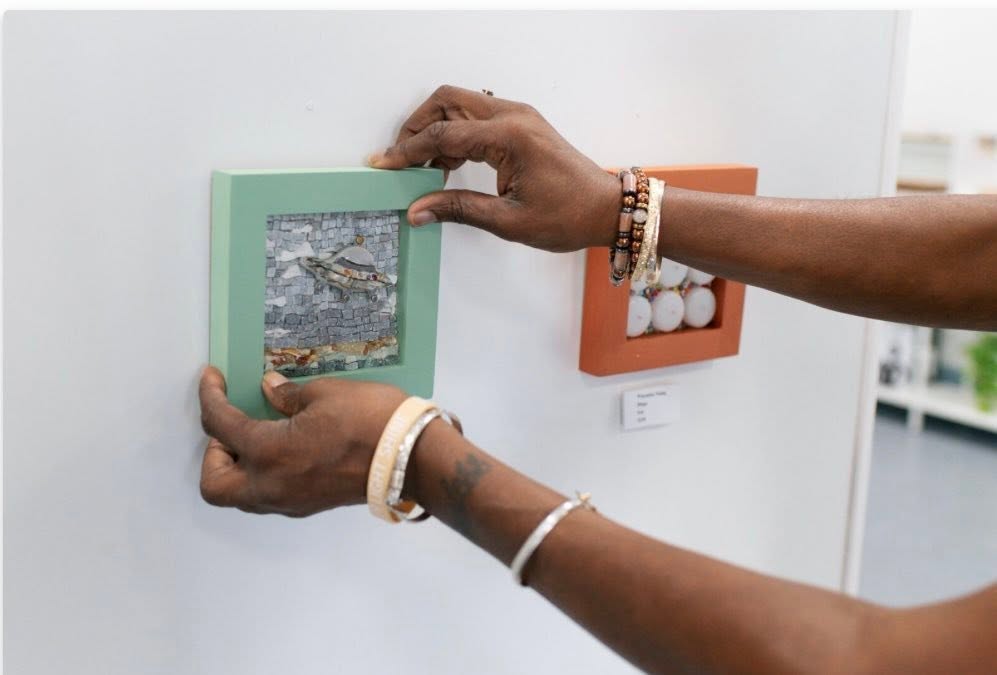 A person with dark skin wearing brass and gold beaded bangles hangs a piece of framed mosaic artwork on a white gallery wall.