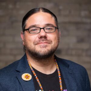 A headshot of a person with light skin, long dark hair pulled back, and dark facial hair wears black rimmed glasses and a blue blazer with a colorful beaded necklace and oval broach featuring a flower.