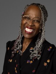 A headshot of a person with dark skin and long gray braids wearing gold rimmed glasses, red lipstick, and a black jacket embellished with flowers smiles enthusiastically.